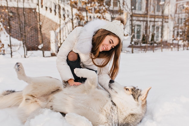 Mattina congelata soleggiata di giovane donna alla moda goduta che gioca con il cane husky nella neve all'aperto. Momenti incantevoli, vere emozioni felici, simpatici animali domestici, vacanze invernali.