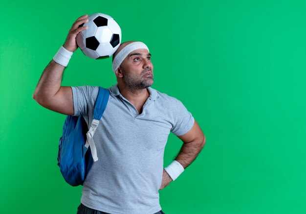 Maturo uomo sportivo in fascia tenendo il pallone da calcio guardando da parte perplesso in piedi sopra la parete verde