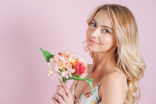 Mazzo biondo sorridente del fiore della tenuta della giovane donna contro il contesto rosa