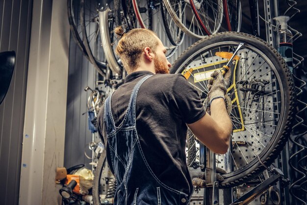 Meccanico barbuto che ripara il pneumatico della ruota di bicicletta in un'officina Vista posteriore, manuale di servizio.