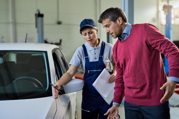 Meccanico femminile e proprietario di auto che comunicano sul guasto del veicolo nell'officina di riparazioni auto