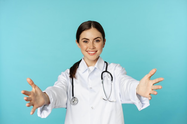 Medico amichevole sorridente, operatore sanitario ragazza, stagista che raggiunge le mani, invitando, abbracciando o ricevendo in braccio, in piedi su sfondo turchese