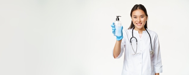 Medico asiatico sorridente che mostra una bottiglia disinfettante per le mani di antisettico che indossa guanti di gomma sterili e c