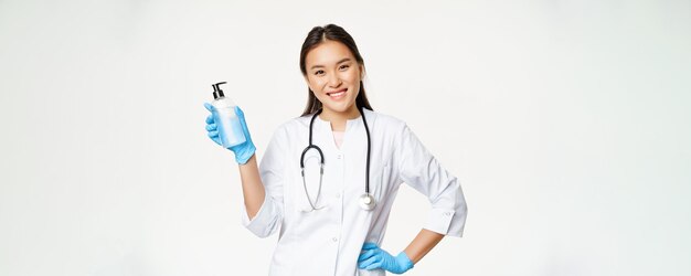 Medico asiatico sorridente che tiene disinfettante per le mani in guanti di gomma che mostrano bottiglia con antisettico per c