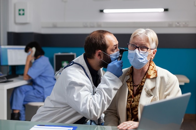 Medico che consulta una donna matura con otoscopio alla visita di controllo, che esegue un esame dell'orecchio per trattare l'infezione durante la pandemia di covid 19. Medico che utilizza lo strumento di otoscopia all'appuntamento di consultazione.