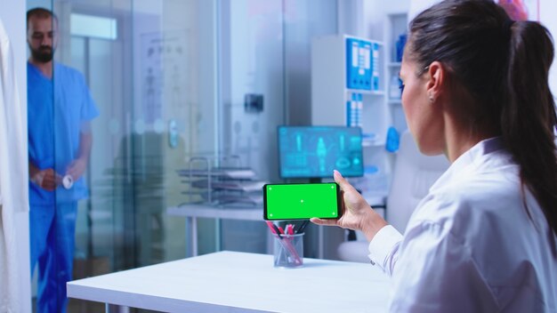 Medico che controlla i risultati del paziente su smartphone con schermo verde nell'armadietto dell'ospedale. L'infermiera in uniforme medica blu chiude la porta di vetro. Specialista sanitario in armadietto ospedaliero utilizzando smartphone con mo