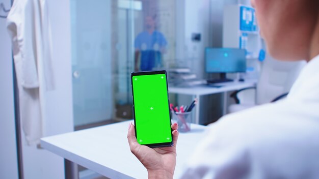 Medico che guarda il telefono con schermo verde nell'armadietto dell'ospedale e infermiere che esce dall'ascensore. Specialista sanitario in armadietto ospedaliero utilizzando smartphone con mockup.