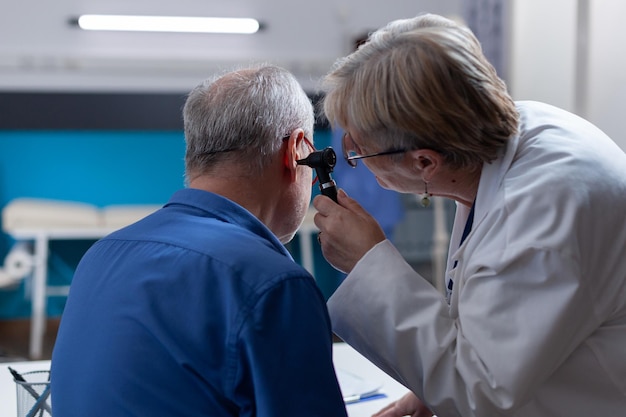 Medico che tiene in mano l'otoscopio per eseguire la consultazione dell'orecchio per il paziente durante la visita di controllo. Medico donna che utilizza uno strumento otologico per esaminare l'infezione e dare consigli medici all'uomo anziano.