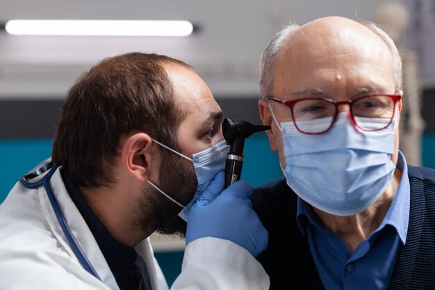 Medico che usa l'otoscopio per fare il controllo dell'orecchio su un uomo anziano, facendo una consultazione otorinolaringoiatrica con il paziente. Medico con maschera facciale che esamina l'infezione per dare un trattamento di prescrizione. Avvicinamento