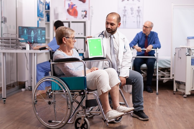 Medico che utilizza tablet pc con schermo verde mentre consulta una donna anziana portatrice di handicap in sedia a rotelle
