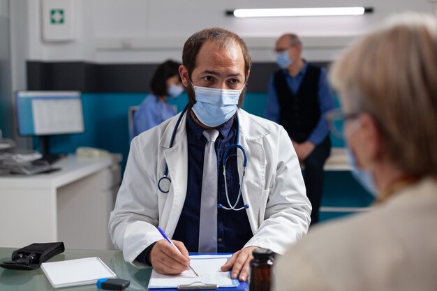 Medico con maschera facciale che prende appunti all'appuntamento di consultazione, parlando con una donna anziana in ufficio. Specialista che parla di medicina e assistenza sanitaria con il paziente durante la pandemia.