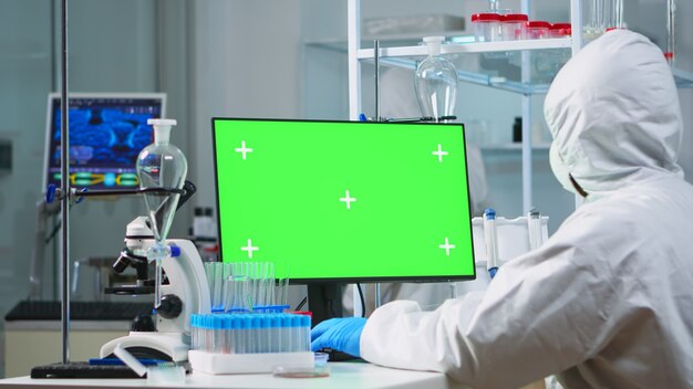 Medico con tuta che lavora al computer con schermo verde in un moderno laboratorio attrezzato. Team di microbiologi che effettuano ricerche sui vaccini scrivendo sul dispositivo con chroma key, isolato, display mockup.