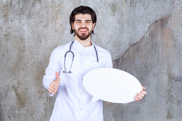 Medico con uno stetoscopio che tiene una scheda informativa vuota ovale e mostra un segno positivo con la mano