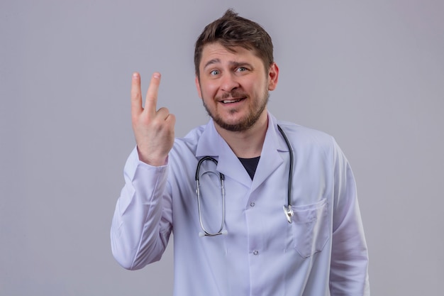 Medico del giovane che indossa le camice e lo stetoscopio che mostrano numero due con le dita con il sorriso sul fronte