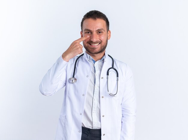 Medico del giovane in camice e con lo stetoscopio intorno al collo sorridente felice e positivo che indica allegramente con il dito indice al suo naso che sta sopra la parete bianca