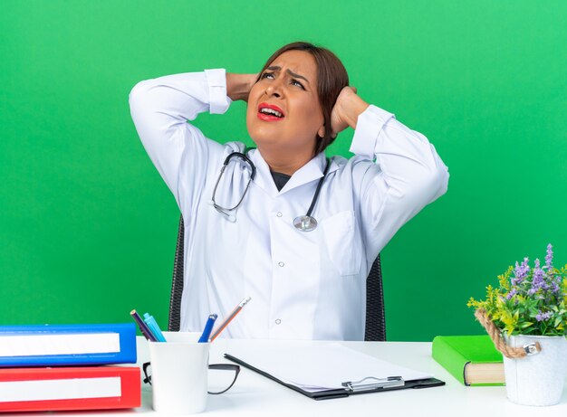 Medico della donna di mezza età in camice bianco con lo stetoscopio che cerca confuso e dispiaciuto seduto al tavolo su green