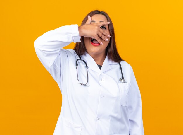 Medico della donna di mezza età in camice bianco con lo stetoscopio coning il viso con i palmi felici e allegri