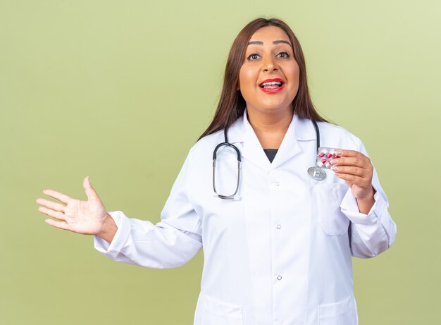 Medico della donna di mezza età in camice bianco con stetoscopio che tiene blister con pillole felice ed eccitato che presenta qualcosa con il braccio della mano in piedi sul verde