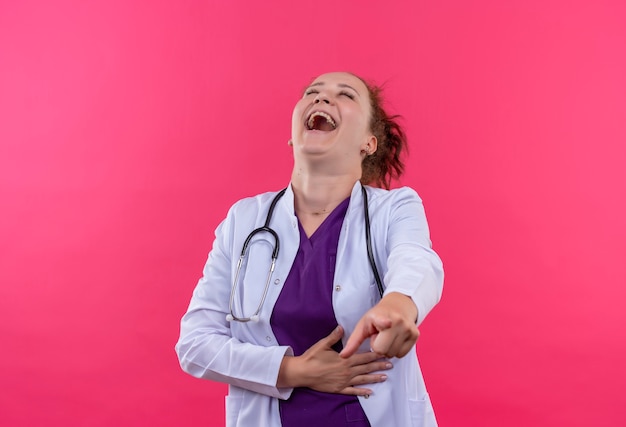 Medico della giovane donna che indossa camice bianco con lo stetoscopio che ride fuori toccando la sua pancia che sta sopra la parete rosa
