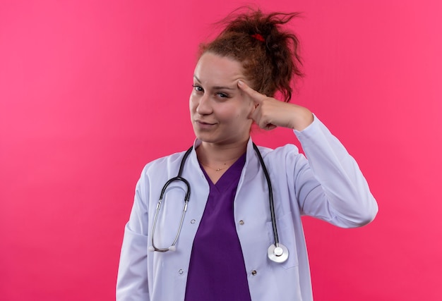 Medico della giovane donna che indossa camice bianco con lo stetoscopio che sembra fiducioso che punta il suo tempio incentrato sul compito in piedi sul muro rosa