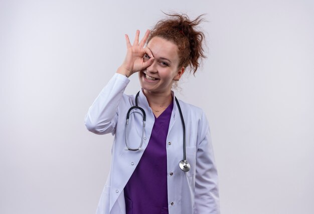 Medico della giovane donna che indossa camice bianco con lo stetoscopio che sorride allegramente facendo segno giusto guardando attraverso questo cartello in piedi sul muro bianco