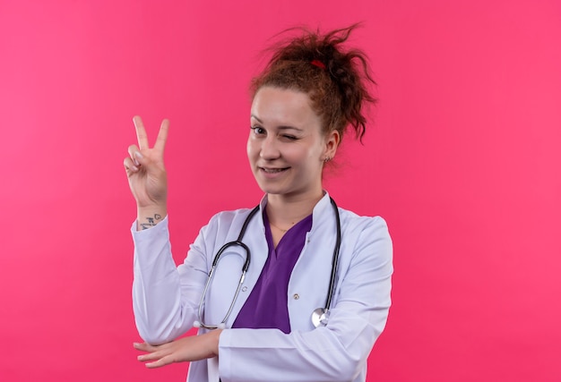 Medico della giovane donna che indossa camice bianco con lo stetoscopio sorridente e ammiccante che mostra il segno di vittoria in piedi sopra il muro rosa