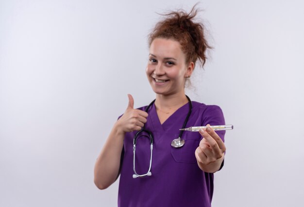 Medico della giovane donna che indossa l'uniforme medica con il termometro della tenuta dello stetoscopio che sorride che mostra i pollici in su che sta sopra il muro bianco