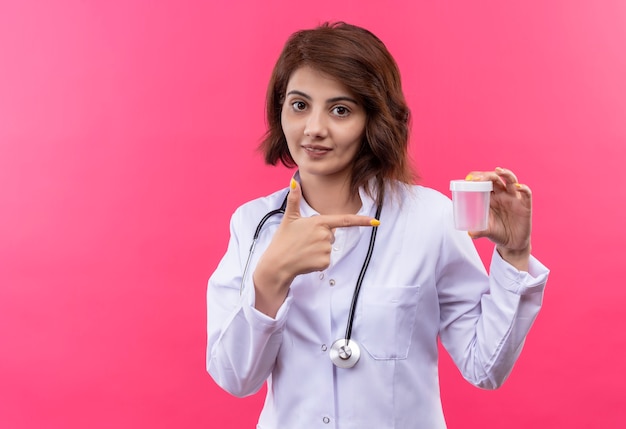 Medico della giovane donna in camice bianco con il dito puntato del barattolo della prova della tenuta dello stetoscopio che sorride fiducioso