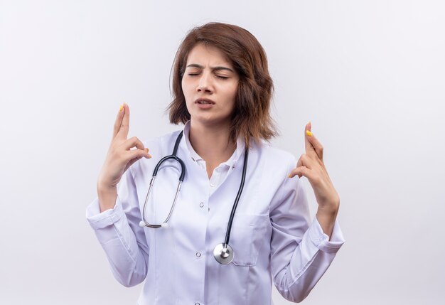 Medico della giovane donna in camice bianco con lo stetoscopio che fa desiderio desiderabile con gli occhi chiusi che attraversano le dita che stanno sopra il muro bianco