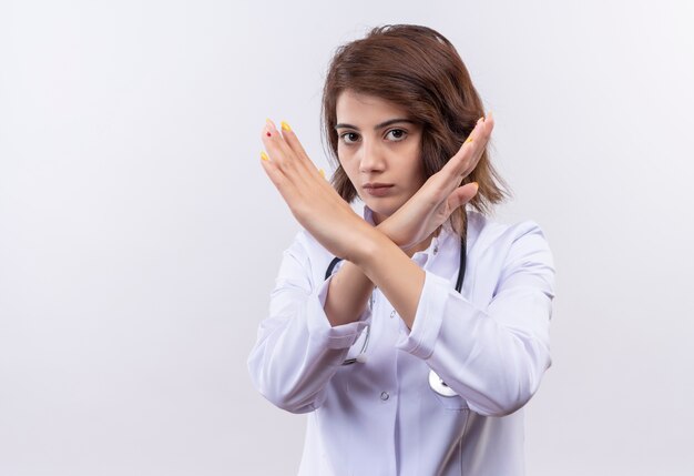 Medico della giovane donna in camice bianco con lo stetoscopio che fa gesto di difesa che attraversa le braccia sul viso in piedi sul muro bianco