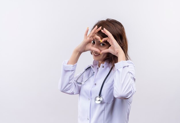 Medico della giovane donna in camice bianco con lo stetoscopio che fa il gesto del cuore con le dita sul viso guardando attraverso le dita sorridenti in piedi sul muro bianco