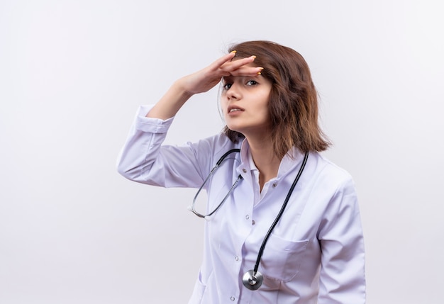 Medico della giovane donna in camice bianco con lo stetoscopio che guarda l'arguzia lontana mano sopra la testa per guardare qualcuno o qualcosa