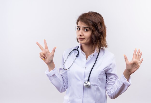 Medico della giovane donna in camice bianco con lo stetoscopio che guarda l'obbiettivo con espressione scettica che mostra e rivolta verso l'alto con le dita numero sette
