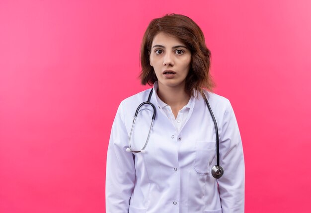 Medico della giovane donna in camice bianco con lo stetoscopio che guarda l'obbiettivo sorpreso e stupito