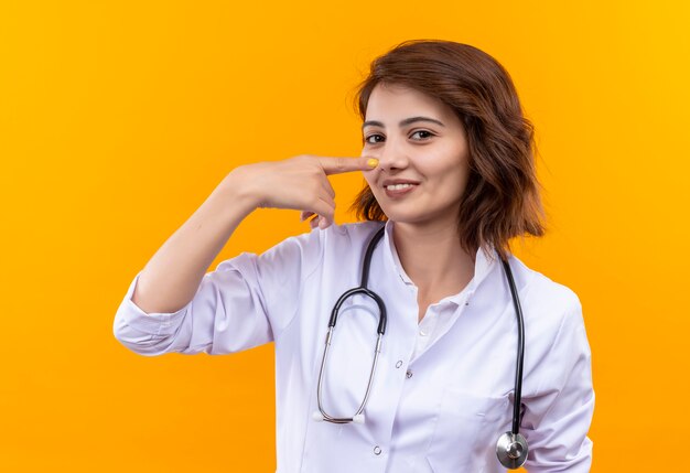 Medico della giovane donna in camice bianco con lo stetoscopio che indica il suo naso sorridente che sta sopra la parete arancione
