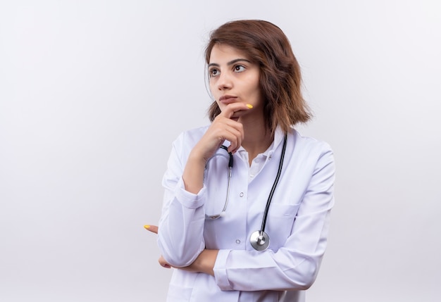 Medico della giovane donna in camice bianco con lo stetoscopio che osserva da parte con la mano sul mento con espressione pensierosa sul pensiero del viso