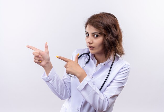 Medico della giovane donna in camice bianco con lo stetoscopio che sembra puntare fiducioso con le dita indice a lato