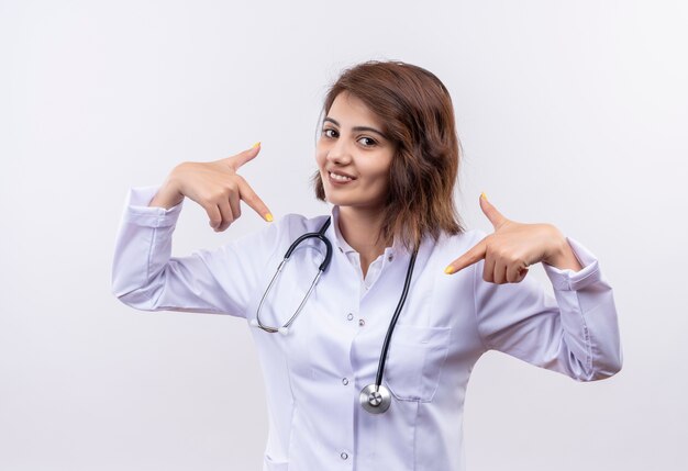 Medico della giovane donna in camice bianco con lo stetoscopio che sembra sorridente fiducioso che indica con le dita a se stessa, soddisfatto di sé e orgoglioso