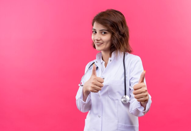 Medico della giovane donna in camice bianco con lo stetoscopio che sorride allegramente che mostra i pollici in su con entrambe le mani che stanno sopra il muro rosa