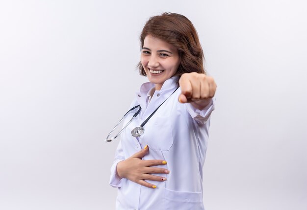 Medico della giovane donna in camice bianco con lo stetoscopio che sorride allegramente indicando con il dito indice che sta sopra il muro bianco