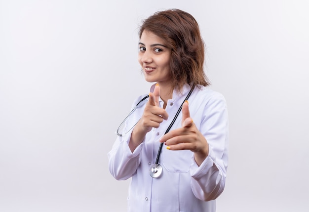 Medico della giovane donna in camice bianco con lo stetoscopio che sorride allegramente indicando con le dita indice con entrambe le mani in piedi sul muro bianco