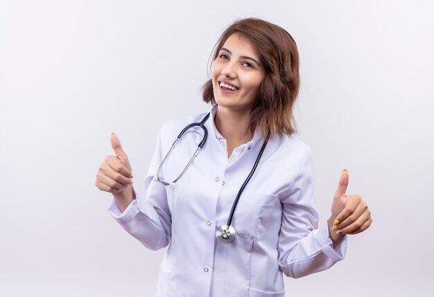 Medico della giovane donna in camice bianco con lo stetoscopio che sorride che mostra i pollici in su con entrambe le mani che stanno sopra il muro bianco