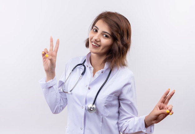 Medico della giovane donna in camice bianco con lo stetoscopio che sorride con la faccia felice che mostra i segni di vittoria che stanno sopra il muro bianco