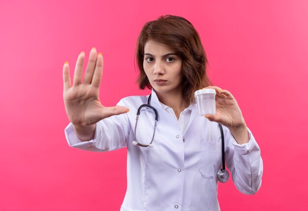 Medico della giovane donna in camice bianco con lo stetoscopio che tiene il barattolo della prova che fa il fanale di arresto con la mano aperta con la faccia seria