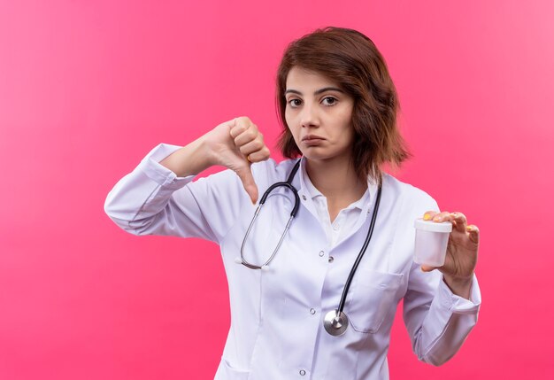 Medico della giovane donna in camice bianco con lo stetoscopio che tiene il barattolo della prova che mostra i pollici giù con il fronte dispiaciuto