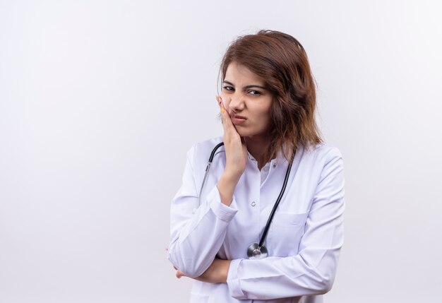 Medico della giovane donna in camice bianco con lo stetoscopio che tocca la guancia con la mano con la faccia accigliata sensazione di dolore in piedi sopra il muro bianco