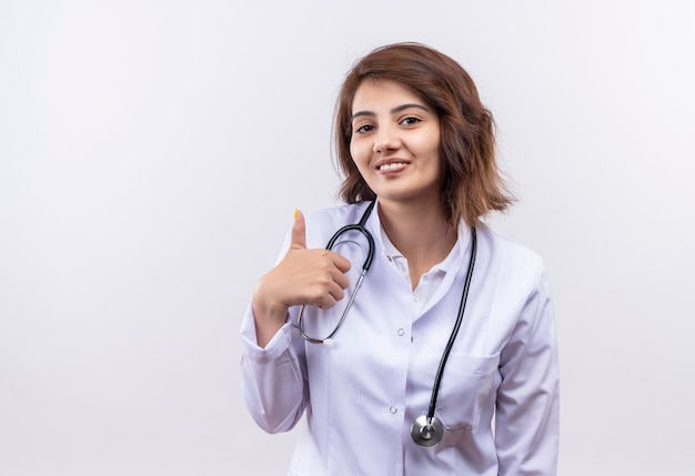 Medico della giovane donna in camice bianco con lo stetoscopio sorridente che mostra i pollici in su che si leva in piedi sopra il wallv bianco
