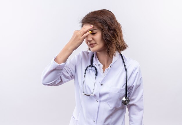 Medico della giovane donna in camice bianco con stetoscopio alla ricerca di pneumatici e naso toccante oberato di lavoro tra gli occhi chiusi