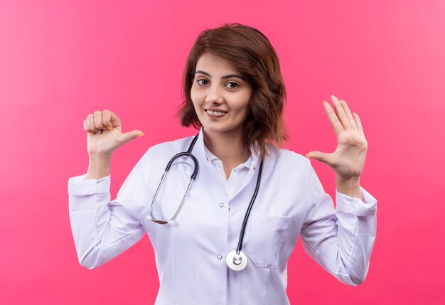 Medico della giovane donna in camice bianco con uno stetoscopio che mostra e rivolto verso l'alto con le dita numero sei sorridente in piedi sul muro rosa
