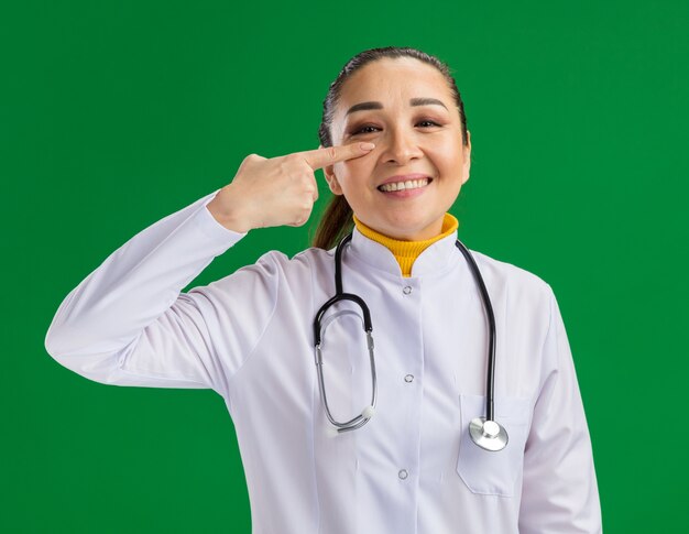 Medico della giovane donna in camice bianco della medicina con lo stetoscopio intorno al collo che indica con il dito indice al suo occhio che sorride allegramente che sta sopra la parete verde